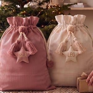 May include: Two large corduroy drawstring sacks in dusty rose and cream colors. Each sack features a decorative star-shaped tag with the names ISLA and LEO printed in gold, accented with wooden beads, tassels, and pom-poms at the top closure.