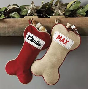 May include: Two dog bone-shaped Christmas stockings. One is red with the name "Chalie" on a white patch, the other is tan with "MAX" on a white patch. Both are hanging from a wooden shelf with star ornaments and greenery.