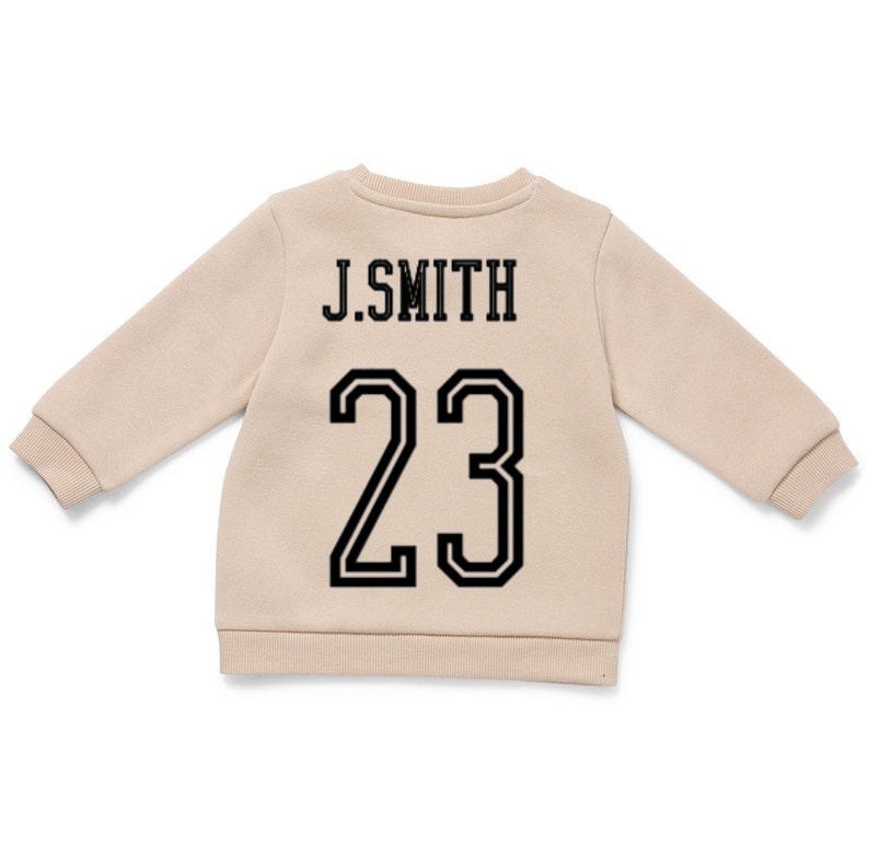 May include: A beige long-sleeve sweatshirt featuring the text J.SMITH in black capital letters above the number 23 in a large black athletic font. The garment has a crew neckline and ribbed cuffs at the sleeves and hem, providing a clean look.