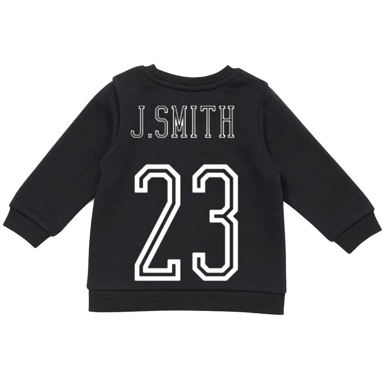 May include: A black long-sleeve sweatshirt featuring the text J.SMITH in white block lettering above the large white number 23. The garment has a ribbed crew neckline, ribbed cuffs, and a ribbed hem, presenting a classic athletic-inspired design.