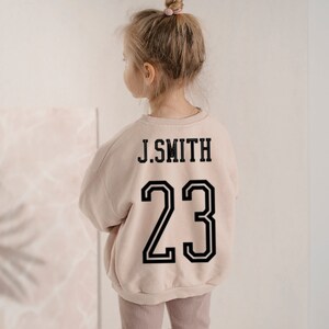 May include: A light beige long-sleeve crewneck sweatshirt featuring the black text J.SMITH and the number 23 printed on the back in a bold athletic font. The garment has a ribbed neckline, cuffs, and hem, providing a structured fit.