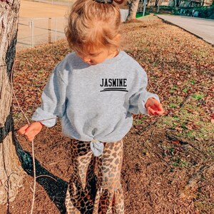 May include: A light gray long-sleeve sweatshirt with a crew neckline and a knotted front hem. The chest features the name JASMINE printed in black capital letters above a horizontal line. Paired with leopard print bell-bottom pants.