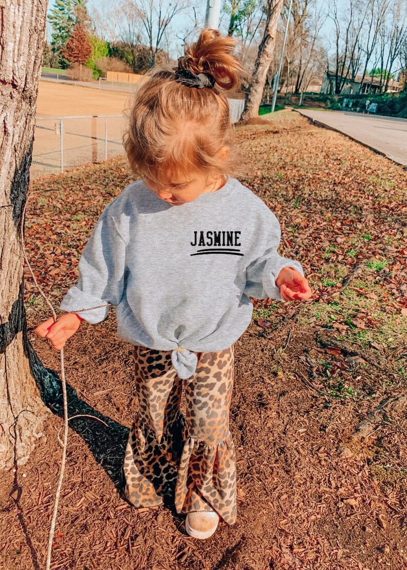 May include: A light gray long-sleeve sweatshirt with a crew neckline and a knotted front hem. The chest features the name JASMINE printed in black capital letters above a horizontal line. Paired with leopard print bell-bottom pants.
