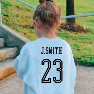 May include: A light gray long-sleeve sweatshirt featuring black block lettering that reads J.SMITH above the large black number 23. The garment has a crew neckline and a standard fit, displaying the back view of the athletic-style jersey design.