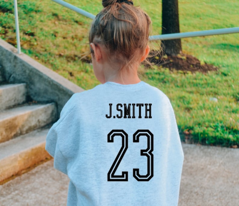 May include: A light gray long-sleeve sweatshirt featuring black block lettering that reads J.SMITH above the large black number 23. The garment has a crew neckline and a standard fit, displaying the back view of the athletic-style jersey design.