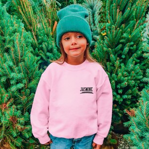 May include: A light pink long-sleeve sweatshirt featuring the name JASMINE printed in black text on the upper left chest, paired with a solid teal knit beanie cap. The garment has a crew neckline and long sleeves with ribbed cuffs.
