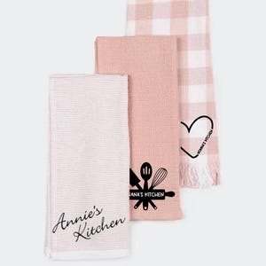 May include: Set of three pink kitchen towels with different designs. One towel has a white and pink gingham pattern, another has a heart with the text "Honnat Kitchen", and the last one has a design of kitchen utensils with the text "Anna's Kitchen" and the text "Annie's Kitchen" in cursive.