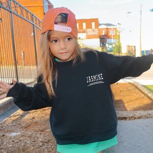May include: A black long-sleeve sweatshirt featuring the name JASMINE printed in white capital letters on the upper left chest, accompanied by two horizontal white lines underneath the text, worn with an orange baseball cap with a pink snapback.