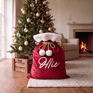 May include: A deep red velvet gift sack featuring a white faux fur trim at the top, a gold braided drawstring with two white pom-poms, and the name Ollie written in white cursive script on the front of the bag for holiday gift storage.
