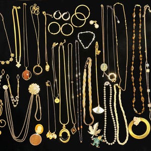 May include: A selection of gold-coloured necklaces, earrings, bracelets, and a brooch displayed on a black surface. The jewellery includes various chain styles, pendants, hoops, and pearl accents. Some pieces feature gemstones and decorative elements.