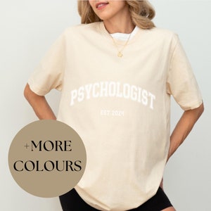 Könnte beinhalten: Ein beigefarbenes Oversize-T-Shirt mit dem Text "PSYCHOLOGIST EST. 2024" in Weiß. Das Shirt wird von einer Person in schwarzen Shorts getragen. Ein brauner Kreis mit dem Text "MEHR FARBEN" befindet sich in der linken unteren Ecke des Bildes.
