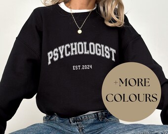 May include: Black sweatshirt with the word "PSYCHOLOGIST" in white arched lettering, with "EST. 2024" below. The sweatshirt has a white collar and is paired with blue jeans and black sneakers. A circular graphic says "+ MORE COLOURS".
