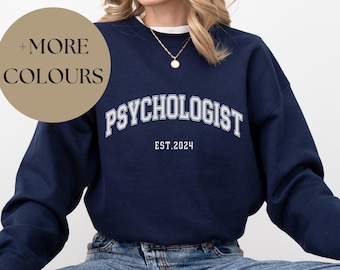 May include: A navy blue sweatshirt with the text "Psychologist Est. 2024" in white lettering. The sweatshirt is being worn by a person sitting down.