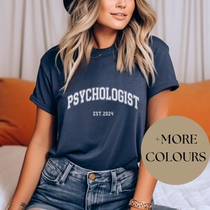 Könnte beinhalten: Ein marineblaues T-Shirt mit dem Text "Psychologist Est. 2024" in Weiß. Das Shirt wird von einer Person mit langen blonden Haaren getragen. Ein brauner Kreis mit dem Text "Mehr Farben" befindet sich in der rechten unteren Ecke des Bildes.