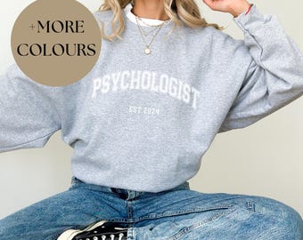 May include: A gray sweatshirt with the text "Psychologist Est. 2024" in white. The model is wearing the sweatshirt with blue jeans and black sneakers.