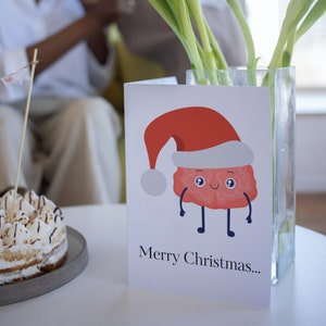 Pu&ograve; includere: Un biglietto bianco con un cervello di cartone animato rosso e bianco che indossa un cappello da Babbo Natale. Il biglietto dice "Buon Natale..."