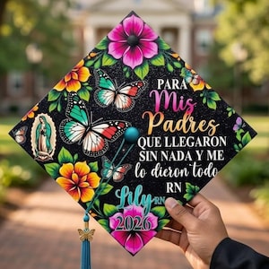 May include: A decorated graduation cap with a black background, featuring colorful flowers, butterflies, and text in Spanish. The cap includes a tassel with a butterfly charm and the name "Lily" and the year "2026".