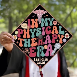 May include: Black graduation cap with a brown border, featuring the words "IN MY PHYSICAL THERAPY ERA" in retro colors. Includes a caduceus, smiley faces, and the name "Isabella" and the year "2026".