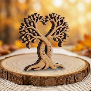 Puede incluir: Escultura de madera de dos árboles entrelazados en forma de corazón. Los nombres "Sarah" y "David" están grabados en los troncos. La escultura se exhibe sobre una rodaja de madera, mostrando una estética natural y rústica.