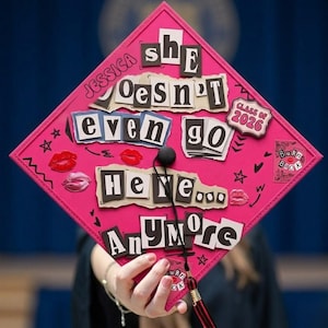 Puede incluir: Una gorra de graduación rosa con el texto "She Doesn't Even Go Here Anymore" en un estilo collage. La gorra también presenta el nombre "Jessica", el año "2026" y gráficos de labios y estrellas.