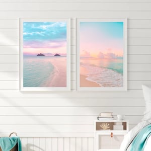May include: Two framed prints of a beach landscape. The prints display a pastel colour scheme of pink, blue, and yellow. The beach scenes show a calm ocean, sandy shore, and a colourful sky. The frames are white, and the overall aesthetic is coastal.