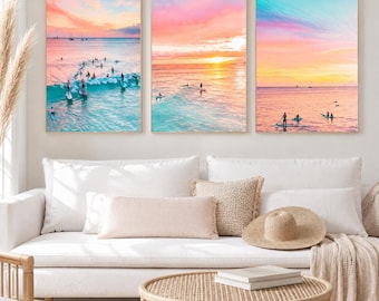 May include: A set of three rectangular wall art prints featuring aerial ocean views at sunset. The images display turquoise water, breaking waves, and people on surfboards and paddleboards under a vibrant pink, orange, and yellow sky.