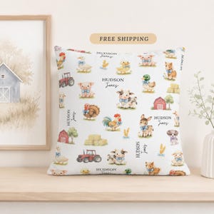 May include: A square white throw pillow featuring a repeating pattern of watercolor farm animals including pigs, cows, ducks, chickens, and donkeys, alongside red tractors, barns, and hay bales. The text "HUDSON James" is printed in black script.