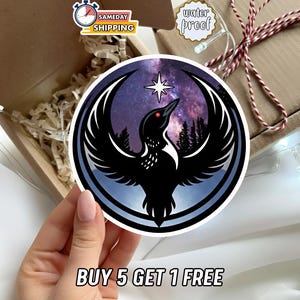 May include: A round sticker featuring a black bird with outstretched wings, a red eye, and a star against a purple and blue galaxy background. The sticker has a white border and the text "BUY 5 GET 1 FREE".