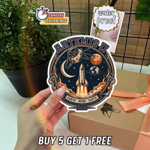 May include: A circular sticker featuring a space-themed design with the text "ARTEMIS II". The design includes a rocket launching, planets, and the text "EARTH MOON EARTH - 2026". The sticker is labeled "water proof".