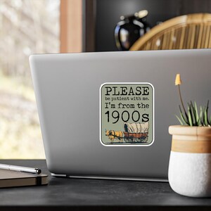 May include: A sticker with a rectangular design featuring the text "PLEASE be patient with me. I'm from the 1900s." Below the text is an illustration of a covered wagon pulled by two oxen. The background is a muted green.