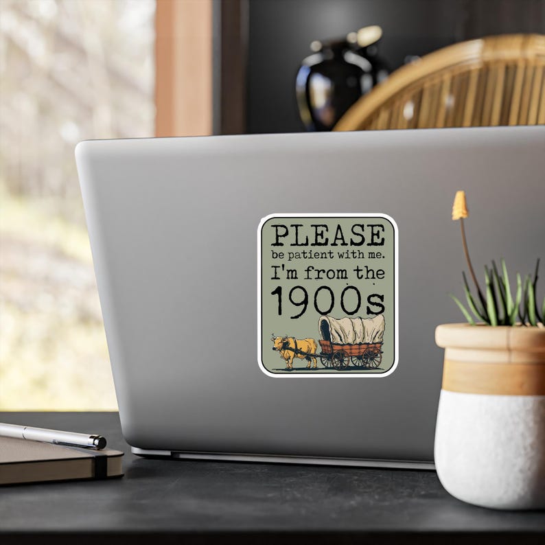 May include: A sticker with a rectangular design featuring the text "PLEASE be patient with me. I'm from the 1900s." Below the text is an illustration of a covered wagon pulled by two oxen. The background is a muted green.