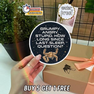 May include: A round sticker with the text "GRUMPY. ANGRY. STUPID. HOW LONG SINCE LAST SLEEP, QUESTION?" and a cartoon robot. The sticker is held in front of a gift box with a peach ribbon. A "water proof" tag is visible.
