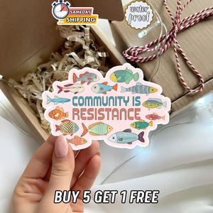 May include: A die-cut sticker with a light pink background featuring various colorful illustrated fish surrounding the text "COMMUNITY IS RESISTANCE" in a bold, dark brown font. The sticker has a white border and a matte finish.