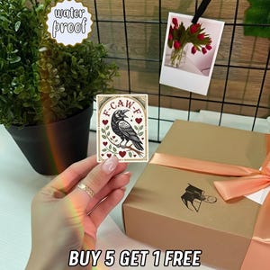 May include: A rectangular sticker featuring a black crow perched on a branch, surrounded by hearts and leaves, with the text "F-CAW-F". The sticker is held in front of a gift box tied with a peach ribbon. The text "water proof" and "BUY 5 GET 1 FREE" are also visible.
