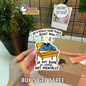 May include: A die-cut sticker featuring a blue hoodie sitting at a wooden school desk with an open book and pencil. A ghost head floats above the hoodie holding a drink. Text reads: IN A REALLY GOOD PLACE RIGHT NOW IN MY BOOK OF COURSE.. NOT MENTALLY.
