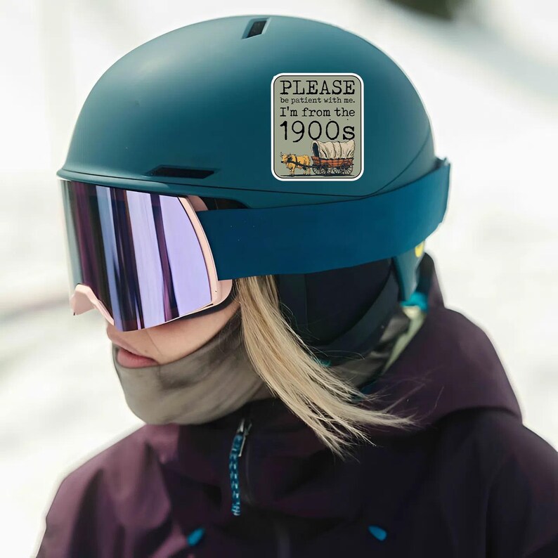 May include: A teal ski helmet with a sticker that reads "PLEASE be patient with me. I'm from the 1900s" and a graphic of a covered wagon. The helmet has a visor with pink and purple lenses.