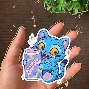 K&ouml;nnte beinhalten: Blauer Katzenaufkleber mit Tigerstreifen, der eine Limonadendose mit der Aufschrift "MY LITTLE SODA POP" h&auml;lt. Die Katze hat gelbe Augen und rosa Akzente. Der Aufkleber ist von Sternen und Glitzer umgeben.