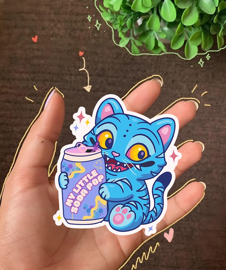 K&ouml;nnte beinhalten: Blauer Katzenaufkleber mit Tigerstreifen, der eine Limonadendose mit der Aufschrift "MY LITTLE SODA POP" h&auml;lt. Die Katze hat gelbe Augen und rosa Akzente. Der Aufkleber ist von Sternen und Glitzer umgeben.