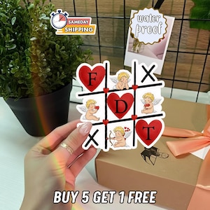 May include: A white sticker with a tic-tac-toe design featuring red hearts and cherubs. The letters "F", "D", and "T" are inside the hearts. The sticker also has the text "BUY 5 GET 1 FREE".