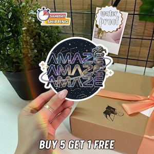May include: A round sticker with the word "AMAZE" repeated three times in a galaxy-themed font. The sticker is held in a hand, with a gift box and a plant in the background. The sticker also has the text "BUY 5 GET 1 FREE".