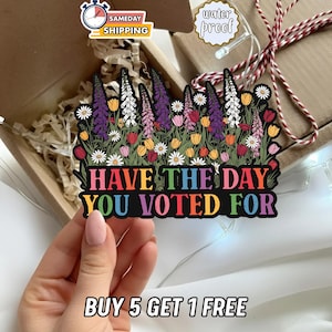 May include: A colorful sticker with the text "HAVE THE DAY YOU VOTED FOR" surrounded by a floral design. The sticker is held by a hand. The image also includes a box and the text "BUY 5 GET 1 FREE".