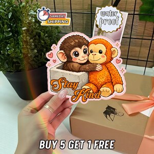 Pode incluir: Um autocolante colorido com dois macacos de desenho animado a abraçar-se. Um é castanho e está num bloco de betão, o outro é laranja. O autocolante tem as palavras "Stay Kind" em fonte laranja. O autocolante é à prova de água.