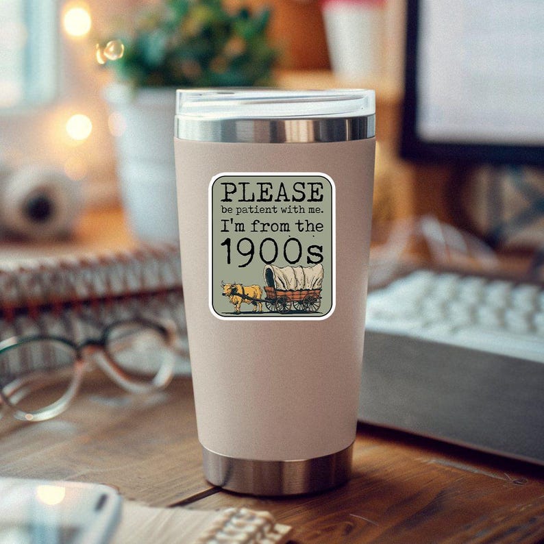 May include: A beige insulated tumbler with a silver lid. A sticker on the tumbler reads "PLEASE be patient with me. I'm from the 1900s" with an illustration of a covered wagon and horses. The tumbler is on a wooden surface.