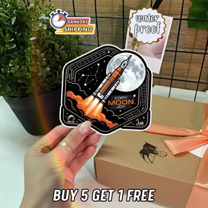 May include: A black sticker with a rocket ship blasting off toward a full moon. The sticker has orange and white flames, a starry background, and the words "COPY MOON JOY". The sticker is held in front of a gift box.
