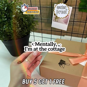 May include: A hand wearing a gold ring is in the foreground. A brown gift box with a peach ribbon is in the background. The text "Mentally, I'm at the cottage" and "BUY 5 GET 1 FREE" are visible. A small plant and a photo with the text "water proof" are also present.