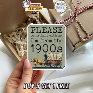 May include: A square sticker with the text "PLEASE be patient with me. I'm from the 1900s" and an illustration of a covered wagon pulled by a bull. The sticker is held in a hand, near a gift box.