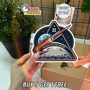 May include: A waterproof sticker featuring a rocket launching from Earth towards the moon. The design includes the text "RETRUN TO THE MOON" and the names of the crew. The sticker is held in front of a gift box.