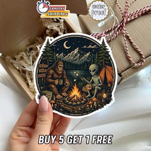 May include: A circular sticker featuring a colorful illustration of a brown Bigfoot and a grey alien sitting by a campfire at night. They are roasting marshmallows in a forest setting with mountains and a crescent moon in the background. A signpost reads Believe, Explore, Discover.