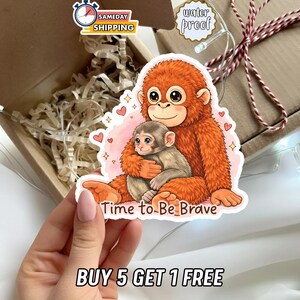 Pode incluir: Um autocolante com um orangotango de desenho animado abraçando um bebê macaco, em um fundo rosa cheio de corações. O texto "Time to Be Brave" é exibido. O autocolante é à prova d'água.