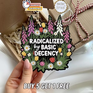 May include: A black sticker with the text "RADICALIZED by BASIC DECENCY" surrounded by colorful flowers. The sticker features pink, white, yellow, and red flowers. The sticker is held in front of a gift box.